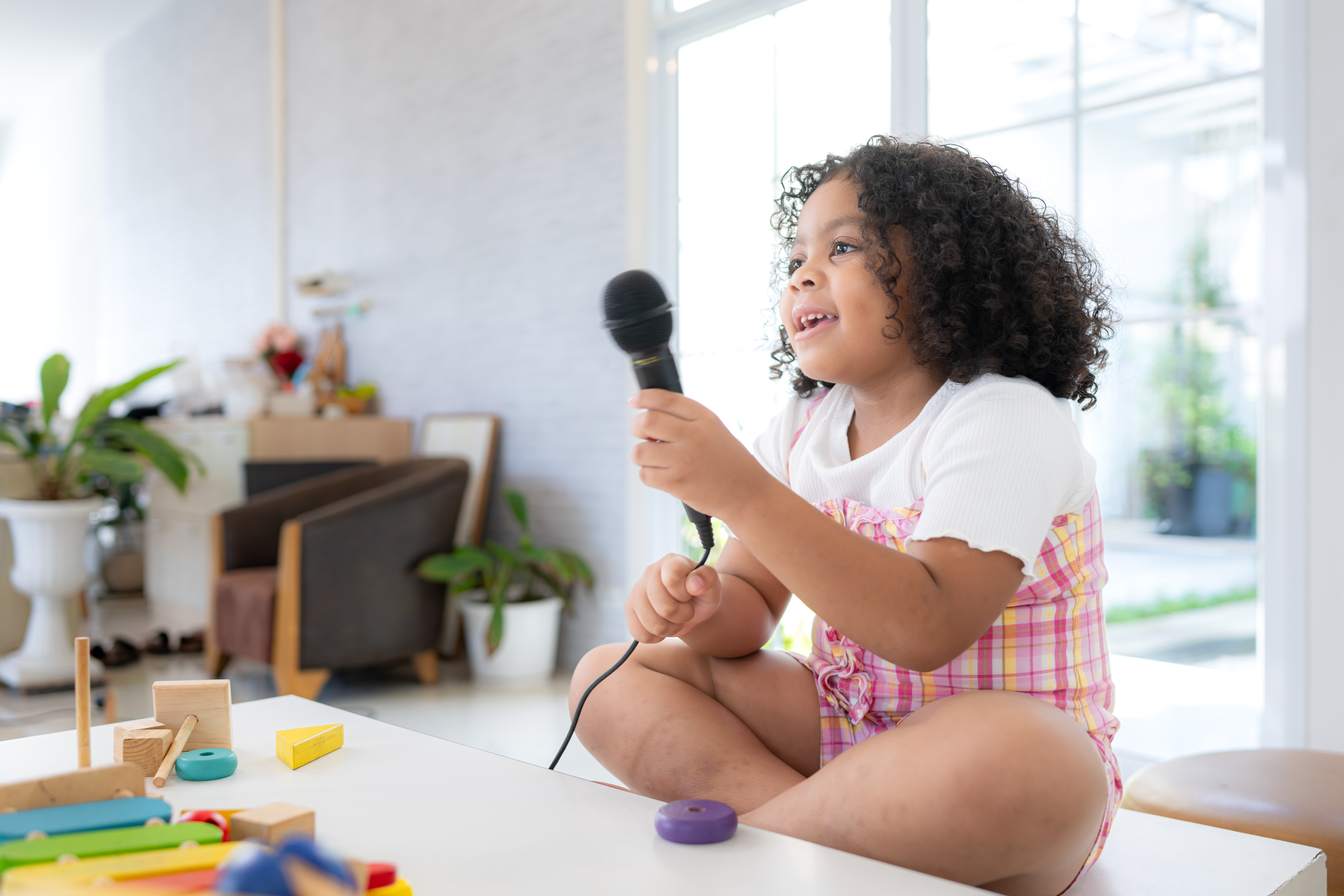 A tiny girl sits on a toy table, clutching a microphone, and sin